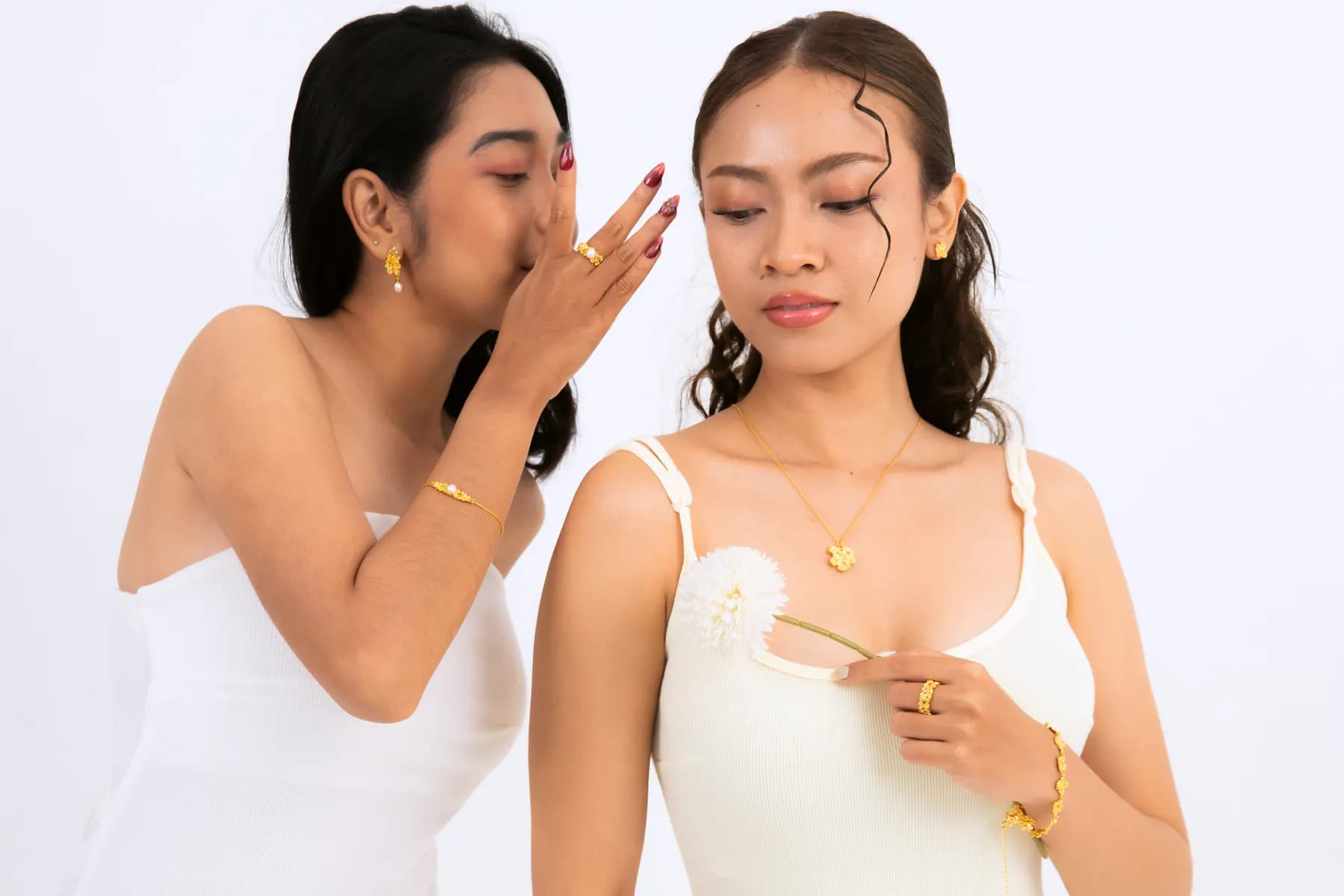 Two models whispering in white, wearing gold pendant necklace, rings and bracelets