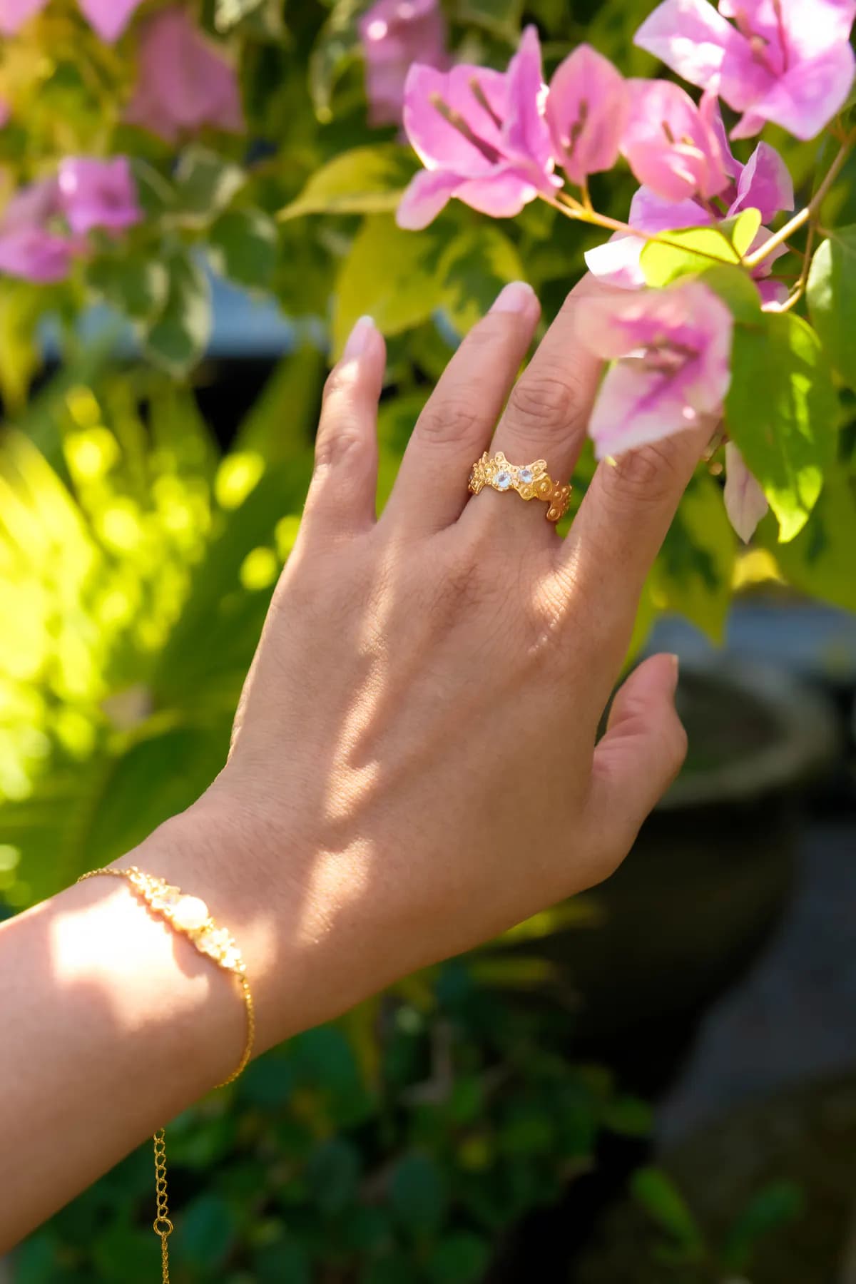 Hand with a gold floral ring and bracelet reaching to pink bougainvillea