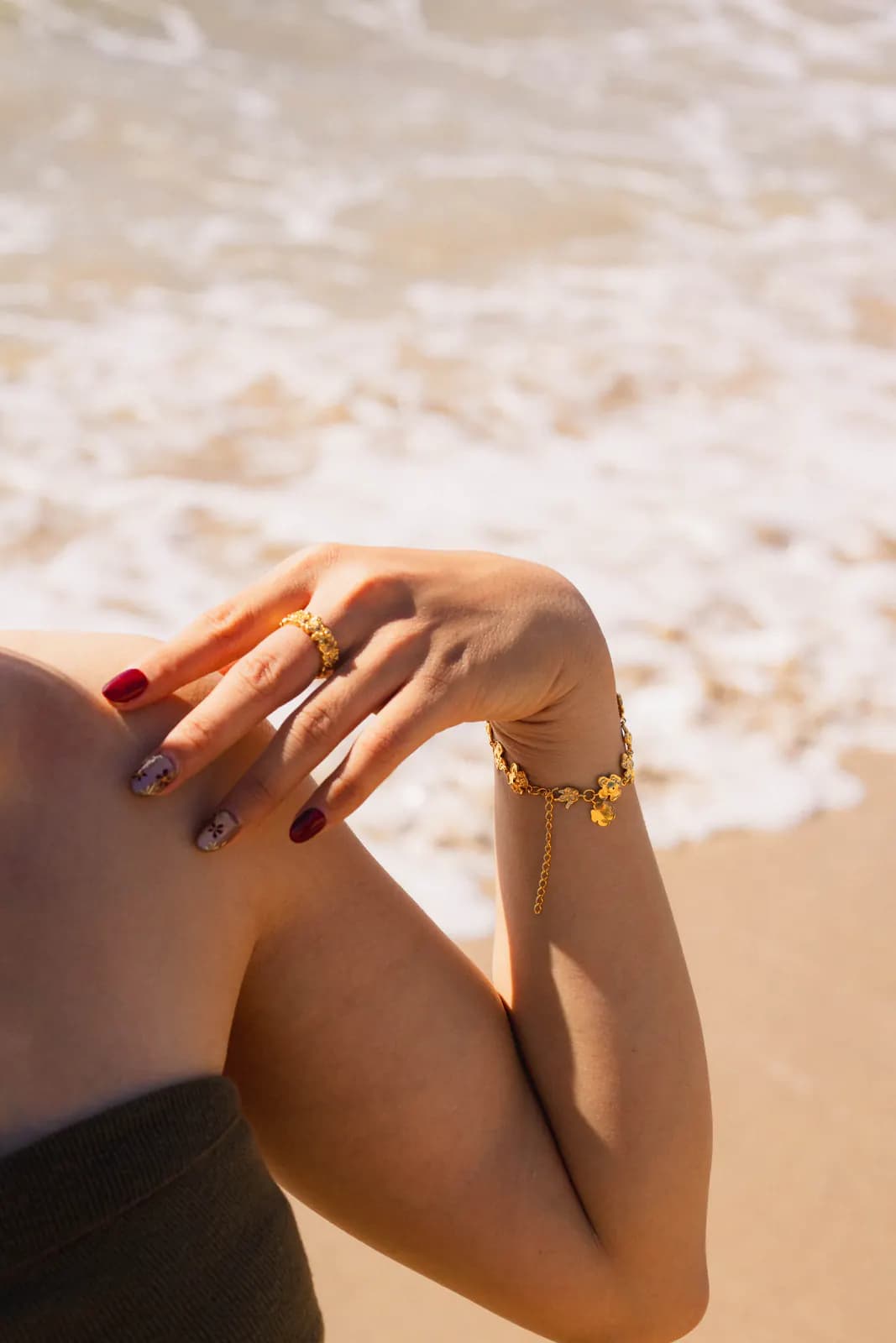 Hand on shoulder wearing a gold ring and floral bracelet against sand and surf