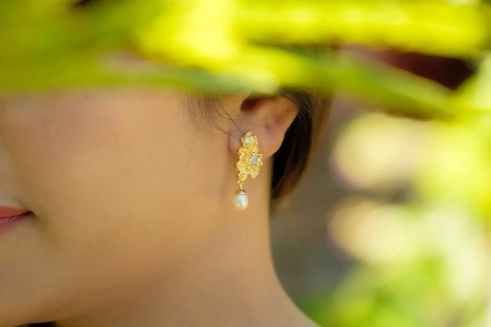 MEDINI gold floral drop earring with a pearl, framed by blurred green leaves