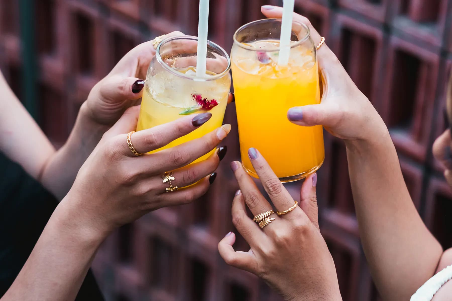 Hands with stacked gold rings clinking orange cocktails against a red lattice wall