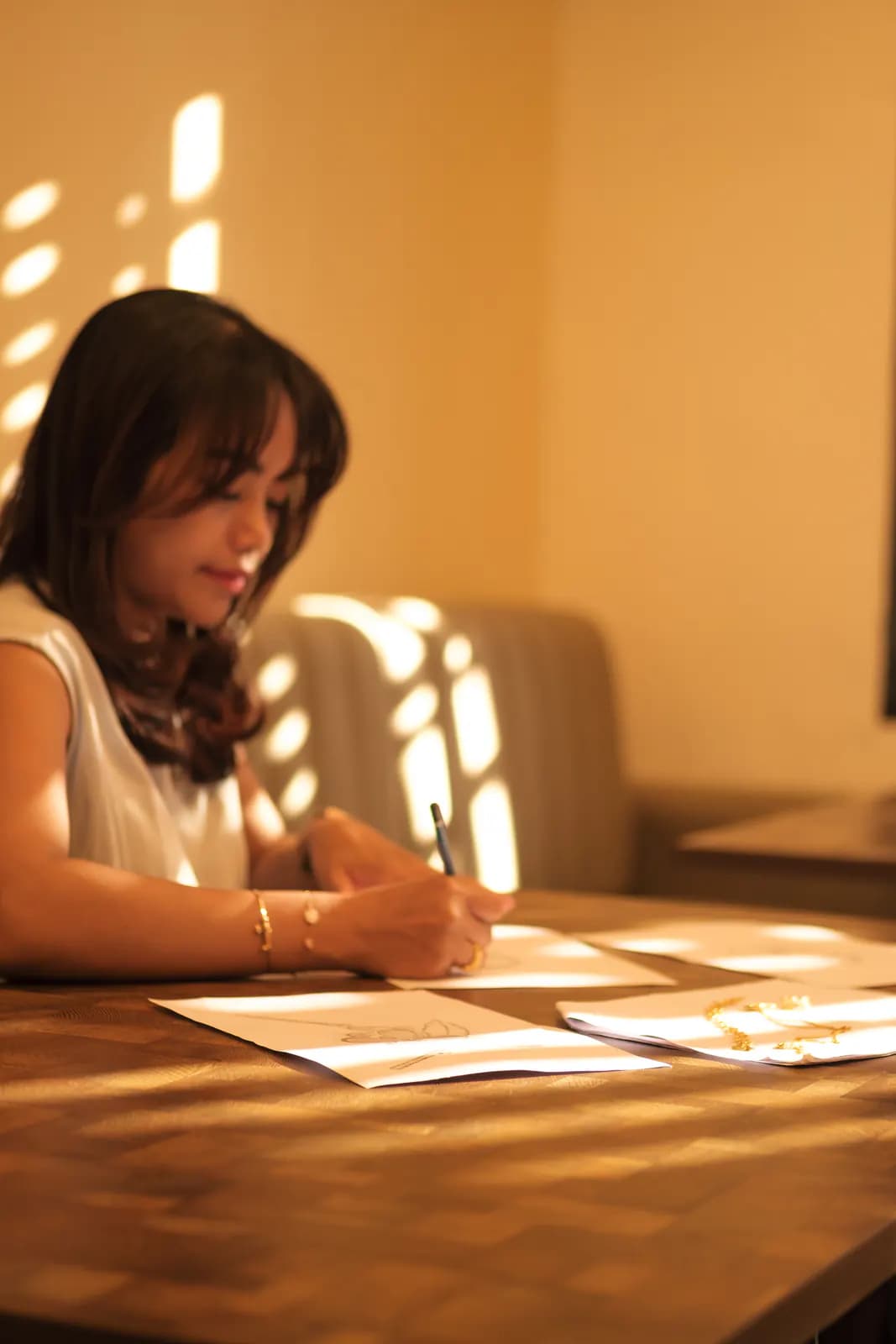 Young woman in white sketching designs at a sunlit wooden table